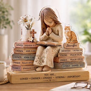May include: A figurine of a girl praying, seated on stacked books with inspirational words and Bible verses. A ginger cat sits nearby. A vase with flowers and a cross are on the left. The base reads "YOU ARE..." and other words.
