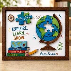 Könnte beinhalten: Ein gerahmtes Wandbild mit einem Holzweltglobus mit Puzzleteil-Ausschnitt, einem Kompass, einem Bücherstapel mit der Aufschrift Atlas, World Facts und Maps, einem Stift und einem Puzzleteil mit dem Text Ms. Ava. Der Text lautet Explore, Learn, Grow.