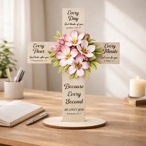 Könnte beinhalten: Holzkreuz mit Blumenschmuck. Das Kreuz trägt die Aufschriften „Jeden Tag“, „Jede Stunde“, „Jede Minute“ und „Denn jede Sekunde“. Es ist mit rosa und weißen Blumen und grünen Blättern verziert. Eine Kerze und ein Buch befinden sich im Hintergrund.