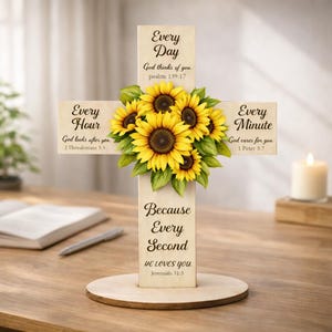 Könnte beinhalten: Ein Holzkreuz mit den Aufschriften „Jeden Tag“, „Jede Stunde“ und „Jede Minute“. Das Kreuz ist mit einem Sonnenblumenstrauß und dem Text „Denn jede Sekunde LIEBT ER DICH“ verziert. Im Hintergrund ist eine brennende Kerze zu sehen.