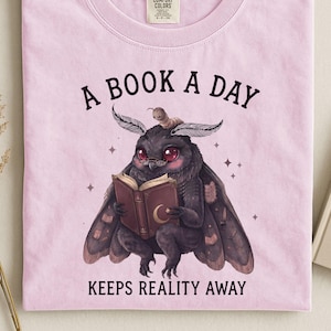 Può includere: Una t-shirt a maniche corte rosa chiaro con la grafica di una falena scura che indossa occhiali e legge un libro. Il testo sopra recita A BOOK A DAY e il testo sotto recita KEEPS REALITY AWAY. La maglietta ha un'etichetta Comfort Colors.