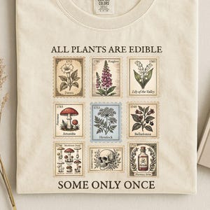 May include: Ivory-colored t-shirt with the text "ALL PLANTS ARE EDIBLE SOME ONLY ONCE." The shirt features nine botanical illustrations resembling postage stamps, including flowers, mushrooms, and a skull. The shirt is made of a soft material.