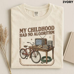 May include: An ivory t-shirt featuring a graphic print with the text 'MY CHILDHOOD HAD NO ALGORITHM'. The illustration depicts a red bicycle, a vintage television, a stack of VHS tapes, a telephone, and a green garden hose on a wooden table.