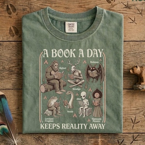 May include: A sage green t-shirt featuring a graphic print with the text A BOOK A DAY KEEPS REALITY AWAY. The illustration depicts mythical creatures including Bigfoot, a Wendigo, Mothman, Loveland Frogman, Nessie, an alien, and a Flatwoods Monster reading books.