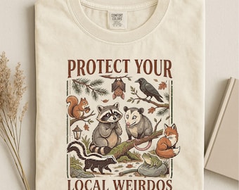 May include: An ivory t-shirt featuring a graphic print with the text "PROTECT YOUR LOCAL WEIRDOS". The illustration depicts a squirrel, a bat, a crow, a raccoon, an opossum, a skunk, a fox, and a frog surrounded by forest elements.