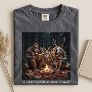 Op de afbeelding: Een antracietgrijs t-shirt met een afbeelding van een Bigfoot, een mot-achtig wezen en een figuur met een hertenkop die in een bos rond een kampvuur zitten te lezen. De tekst onder de afbeelding luidt, A BOOK A DAY KEEPS REALITY AWAY.