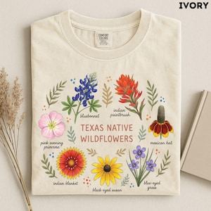 May include: An ivory t-shirt featuring a botanical illustration of Texas native wildflowers including bluebonnet, indian paintbrush, pink evening primrose, mexican hat, indian blanket, black-eyed susan, and blue-eyed grass with corresponding text labels.