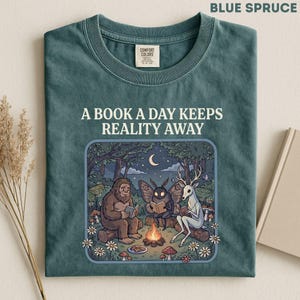 May include: A blue spruce t-shirt featuring the text A BOOK A DAY KEEPS REALITY AWAY above a graphic of a sasquatch, a moth, and a deer sitting around a campfire in a forest at night, each reading a book under a crescent moon and stars.