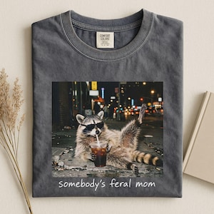 Pode incluir: Uma t-shirt de gola redonda cinzento-carvão com um gráfico de um guaxinim a usar óculos de sol pretos, a segurar um copo de plástico com uma palhinha e sentado numa rua à noite. O texto "somebody's feral mom" está impresso abaixo do gráfico.