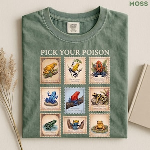 Könnte beinhalten: Ein moosgrünes T-Shirt mit einem Grafikdruck von neun briefmarkenartigen Illustrationen verschiedener Pfeilgiftfrösche. Der Text "PICK YOUR POISON" steht über dem Raster, wobei jeder Frosch mit seinem Namen beschriftet ist.