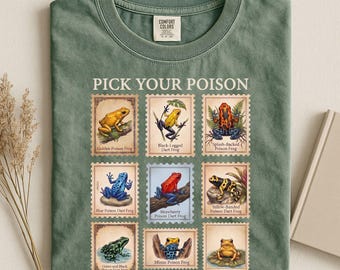 May include: A moss green t-shirt featuring a graphic print of nine postage stamp-style illustrations of various poison dart frogs. The text "PICK YOUR POISON" is printed above the grid of frogs, with each frog labelled by its specific name.