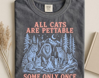 May include: A charcoal grey t-shirt featuring a graphic print of various wild cats around a campfire in a forest setting. The text reads ALL CATS ARE PETTABLE at the top and SOME ONLY ONCE at the bottom in a light pink, distressed font style.