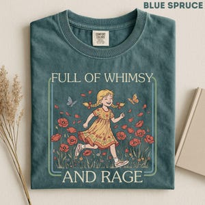 Puede incluir: Una camiseta color abeto azul con un gráfico de una persona con trenzas rubias y vestido amarillo corriendo entre amapolas rojas. El texto dice FULL OF WHIMSY AND RAGE. La camiseta tiene cuello redondo y una etiqueta de Comfort Colors en el cuello.