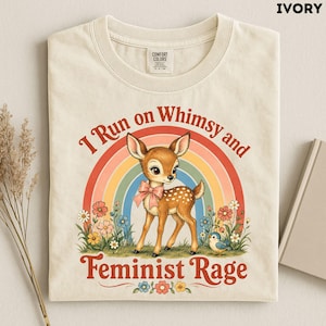 May include: An ivory t-shirt featuring a graphic of a fawn wearing a pink bow, standing in front of a rainbow and flowers. The text reads I Run on Whimsy and Feminist Rage. The shirt has a crew neckline and a Comfort Colors brand label inside.