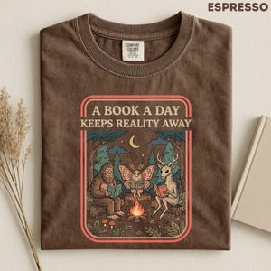 May include: A dark brown t-shirt featuring a graphic print of a sasquatch, a moth, and a deer sitting around a campfire reading books. The text above the illustration reads A BOOK A DAY KEEPS REALITY AWAY. The shirt has a crew neckline and short sleeves.