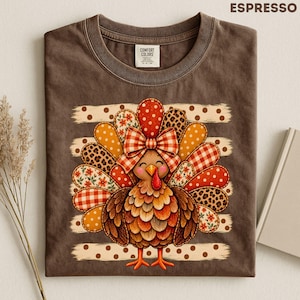 Pode incluir: Uma t-shirt castanho espresso com o gráfico de um peru com um grande laço aos quadrados. As penas da cauda do peru estão decoradas com vários padrões, incluindo bolinhas, estampado de leopardo, xadrez vichy e flores. O texto ESPRESSO é visível.