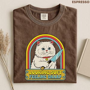 Puede incluir: Camiseta color espresso con un gato de dibujos animados sosteniendo un cuchillo dentro de un arco iris. El gato tiene ojos azules y un collar rojo con una campana. El texto "LOOKING SOFT FEELING SHARP" está debajo.