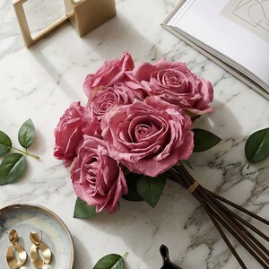 May include: A bouquet of mauve roses with green leaves, tied with a brown rubber band. The roses are arranged on a white marble surface, with a gold-colored frame and a decorative plate with gold earrings.