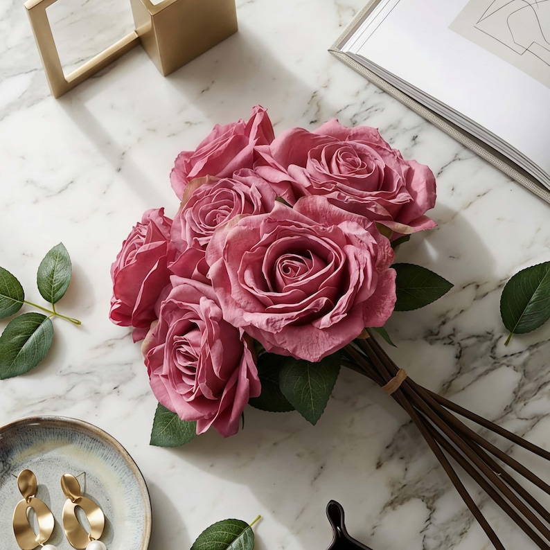 May include: A bouquet of mauve roses with green leaves, tied with a brown rubber band. The roses are arranged on a white marble surface, with a gold-colored frame and a decorative plate with gold earrings.