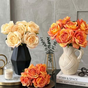 Könnte beinhalten: Arrangement künstlicher Rosen in verschiedenen Vasen. Die Rosen sind in Pfirsich- und Orangetönen gehalten. Eine schwarze geometrische Vase, eine weiße strukturierte Vase und ein goldfarbener Teller halten die Blumen. Dekorative Artikel: Kerze und Bücher.