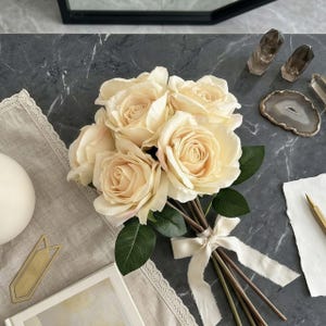 May include: A bouquet of cream-colored roses tied with a cream ribbon. The roses have green leaves and are arranged on a gray marble surface. Other items include crystals, a decorative coaster, and a pen.