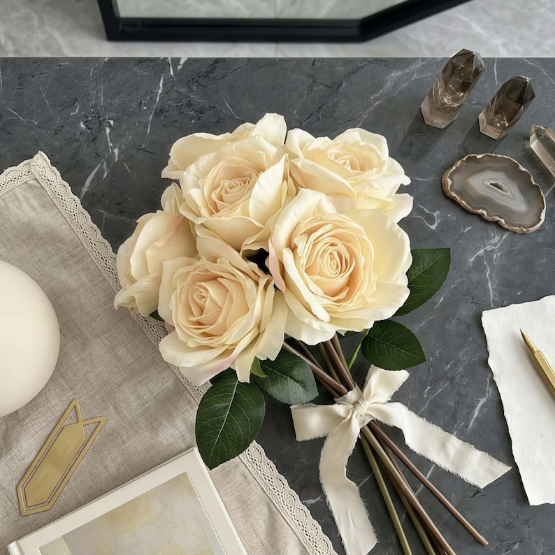 May include: A bouquet of cream-colored roses tied with a cream ribbon. The roses have green leaves and are arranged on a gray marble surface. Other items include crystals, a decorative coaster, and a pen.