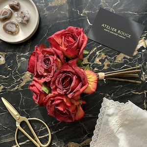 May include: A bouquet of red roses with yellow edges, tied with twine, rests on a black marble surface. Gold scissors, a white plate with stones, and a card with the text "ATELIER ROUGE HAUTE COUTURE FLORALE" are also present.