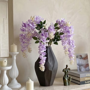 Puede incluir: Un jarrón geométrico marrón oscuro contiene un arreglo de glicinas artificiales. Las flores en cascada son una mezcla de morado y blanco, con hojas verdes. Candelabros y velas blancas a la izquierda, con una pequeña escultura de bronce y libros a la derecha.