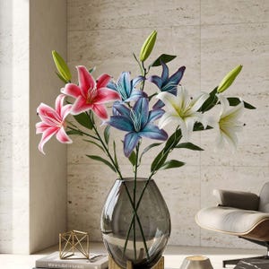 Könnte beinhalten: Ein Strauß künstlicher Lilien in einer grauen Glasvase. Die Lilien sind in Rosa-, Blau- und Weißtönen gehalten, mit grünen Stielen und Blättern. Die Vase steht auf einem goldfarbenen Sockel. Der Hintergrund ist eine neutral getönte Wand.