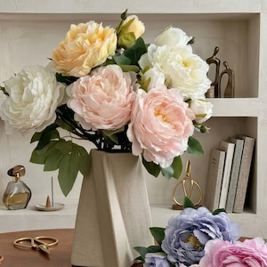 Peut inclure: Un bouquet de pivoines artificielles dans un vase géométrique beige. Les fleurs sont dans des tons de blanc, pêche et rose, avec des feuilles vertes. Le vase est posé sur une table en bois, avec d'autres objets décoratifs en arrière-plan.