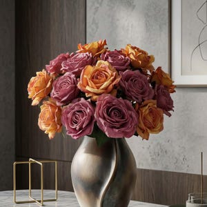 May include: A bouquet of roses in shades of orange and mauve fills a metallic vase. The flowers are in full bloom, showcasing their layered petals. The vase has a unique, curved design. The arrangement sits on a marble surface.