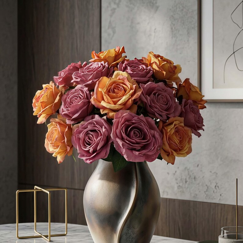 May include: A bouquet of roses in shades of orange and mauve fills a metallic vase. The flowers are in full bloom, showcasing their layered petals. The vase has a unique, curved design. The arrangement sits on a marble surface.