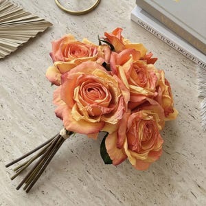 May include: A bouquet of orange and yellow roses tied together with twine. The roses have multiple layers of petals and are arranged with green stems. The bouquet is set on a light-colored, textured surface.