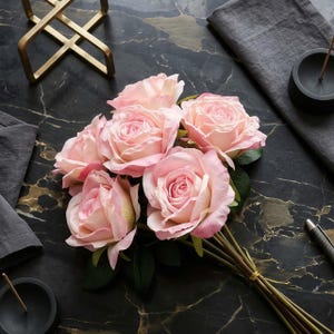 May include: A bouquet of six light pink roses with green leaves, tied with a gold string. The roses are arranged on a black marble surface with gold veining, alongside a gold geometric sculpture and a gray napkin.