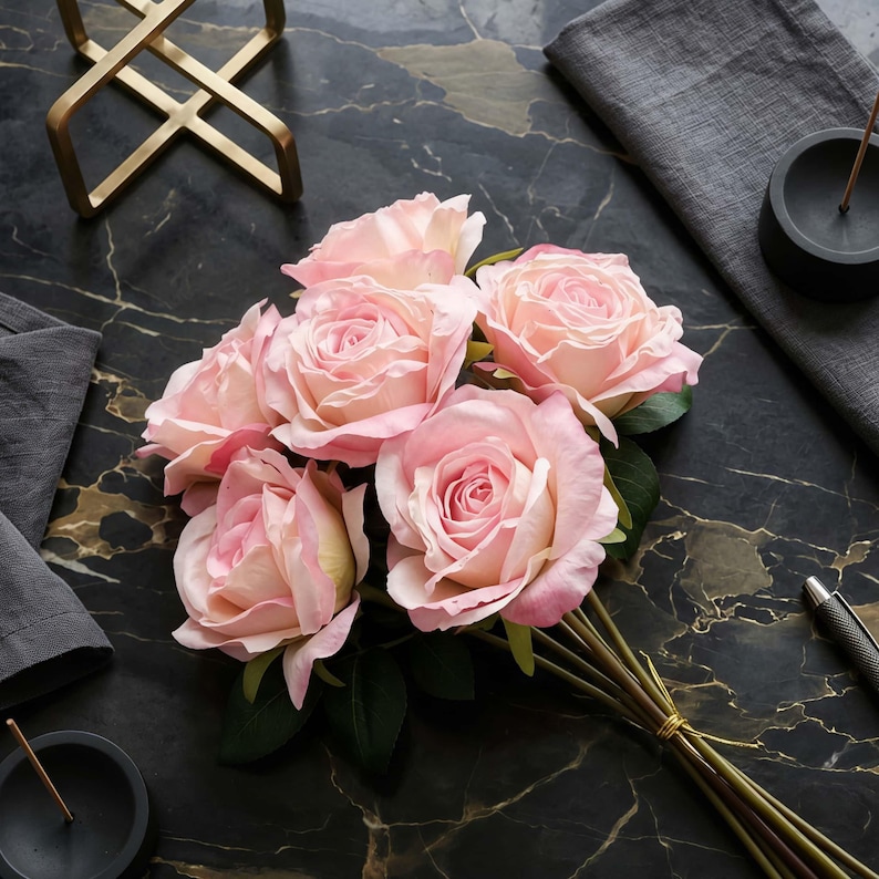 May include: A bouquet of six light pink roses with green leaves, tied with a gold string. The roses are arranged on a black marble surface with gold veining, alongside a gold geometric sculpture and a gray napkin.