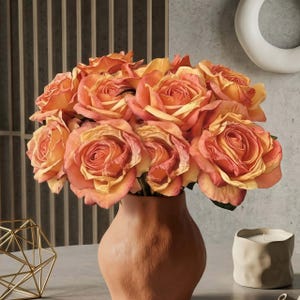 May include: A bouquet of orange and yellow roses in a terracotta vase. The roses have multiple layers of petals and are arranged in a full, lush display. The vase is a warm, earthy tone, and the background is a neutral gray.