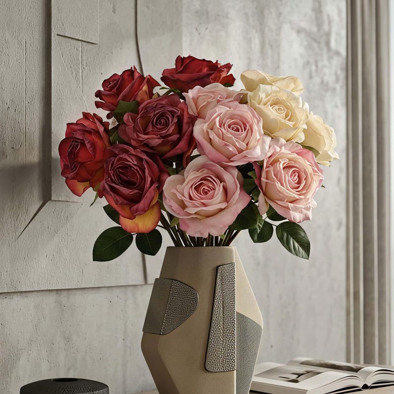 May include: A bouquet of roses in shades of red, pink, and cream, arranged in a geometric vase with textured accents. The flowers have green leaves and are set against a neutral background, creating a sophisticated floral arrangement.