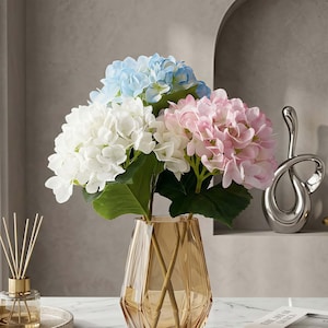 Puede incluir: Un ramo de hortensias artificiales en un jarrón decorativo de cristal ámbar. Las flores son blancas, azul claro y rosas, con hojas verdes. El jarrón tiene un diseño geométrico. Una escultura plateada y un difusor de caña están en el fondo.