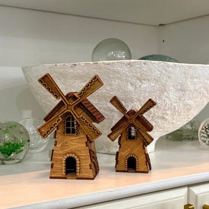 May include: Two wooden windmill figurines with intricate details. The windmills have brown blades, red roofs, and arched doorways. They are displayed on a white shelf, alongside decorative glass spheres and a large white bowl.
