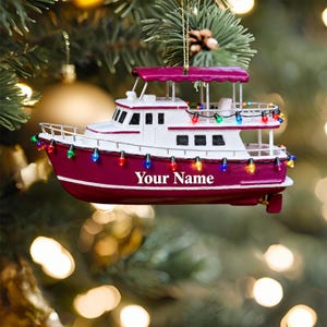May include: A burgundy and white boat-shaped Christmas ornament with colourful lights. The ornament has the text "Your Name" on the side. The boat is hanging from a gold string in front of a blurred background of Christmas tree branches and lights.
