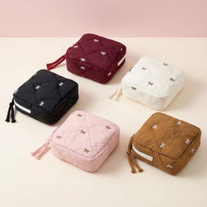 May include: Five square quilted pouches in black, burgundy, white, pink, and brown. Each pouch has a bow pattern and a zip closure. The pouches have a braided handle.