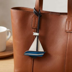 May include: A brown leather tote bag with a sailboat charm. The charm is white, blue, and black, and is attached to the bag's handle with a black leather strap. The bag is a medium size and has a simple design.