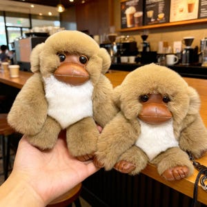 Op de afbeelding: Twee pluche speelgoedvogelbekdieren. Het speelgoed heeft bruine vacht, witte buiken en bruine, leerachtige snavels en voeten. De vogelbekdieren hebben zwarte ogen en zitten op een houten oppervlak.