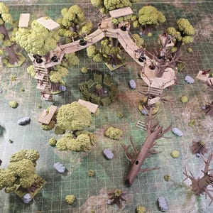 May include: A tabletop gaming terrain set featuring multiple miniature trees with green foliage, wooden platforms, and connecting rope bridges. The scene includes several brown tree trunks, small gray rocks, and a green grid-patterned play mat.