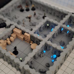 Pode incluir: Um conjunto de terreno de masmorra cinzento de várias salas com figuras em miniatura, barris, caixas e uma estante. A masmorra tem detalhes azuis brilhantes e é projetada para jogos de RPG de mesa.