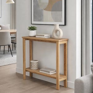 Può includere: Una consolle in legno chiaro con un ripiano inferiore, con libri e un contenitore decorativo. Un'opera d'arte astratta incorniciata è appesa sopra. Un vaso bianco e una pianta sono sul tavolo. La stanza ha toni neutri.