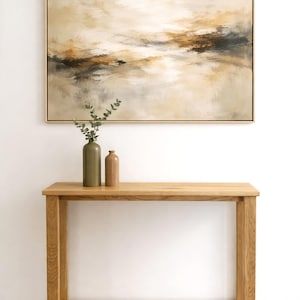 Puede incluir: Una pintura abstracta de paisaje en un marco dorado cuelga sobre una consola de madera. Dos jarrones, uno verde oliva y otro marrón, están sobre la mesa. La pintura presenta tonos neutros de beige, marrón y dorado.