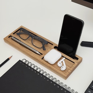 Pode incluir: Um organizador de mesa de madeira com compartimentos para smartphone, óculos, caneta e fones de ouvido sem fio. Um caderno preto, um lápis e um teclado também são visíveis.