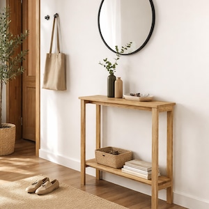 Puede incluir: Una consola de madera clara con un estante inferior, que contiene una cesta tejida y libros. Un espejo redondo con marco negro cuelga encima, con jarrones decorativos y una pequeña bandeja. Una bolsa tote beige cuelga de la pared.