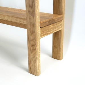 May include: A close-up of a wooden shelf with a light brown finish. The shelf has a rectangular design with a lower shelf and two supporting legs. The wood grain is visible, and the piece is set against a white background.